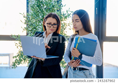 Teacher showing laptop screen to student holding folders near window in modern office Teacher showing laptop screen to student holding folders near window in modern office 143287902
