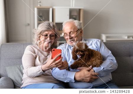 Senior couple checking smartphone relaxing on sofa with poodle dog Senior couple checking smartphone relaxing on sofa with poodle dog 143289428
