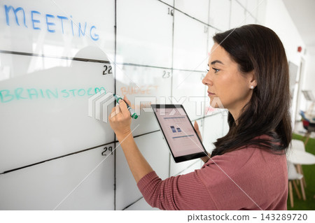 Professional woman writing on large whiteboard calendar while holding tablet Professional woman writing on large whiteboard calendar while holding tablet 143289720