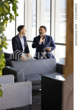 Two businessmen sitting in office lounge, discussing information during workday Two businessmen sitting in office lounge, discussing information during workday 143289829