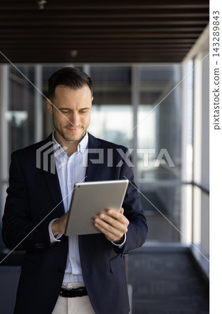 Focused businessman using digital tablet stands alone in office hallway Focused businessman using digital tablet stands alone in office hallway 143289843