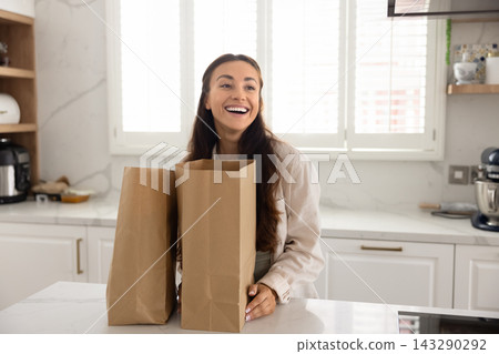 Happy young woman standing in kitchen with two grocery bags Happy young woman standing in kitchen with two grocery bags 143290292