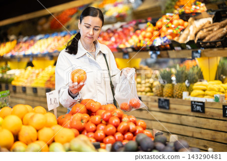 Woman carefully selects fresh tomatoes, standing near the counter in supermarket Woman carefully selects fresh tomatoes, standing near the counter in supermarket 143290481