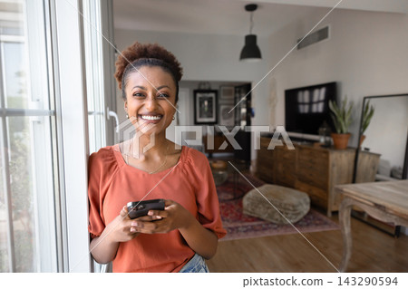 Smiling woman using smartphone standing by window at home 143290594