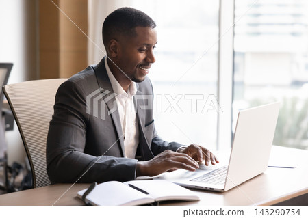 Smiling Black businessman working on digital business tasks using laptop Smiling Black businessman working on digital business tasks using laptop 143290754
