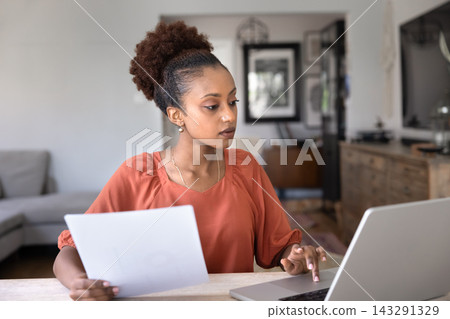 African woman analyzing documents seated at desk with laptop African woman analyzing documents seated at desk with laptop 143291329