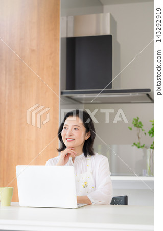 一位微笑的五十多歲女性在家裡的廚房裡一邊使用電腦一邊打字,擺出一副若有所思、憂心忡忡的姿態。 一位微笑的五十多歲女性在家裡的廚房裡一邊使用電腦一邊打字,擺出一副若有所思、憂心忡忡的姿態。 143292919