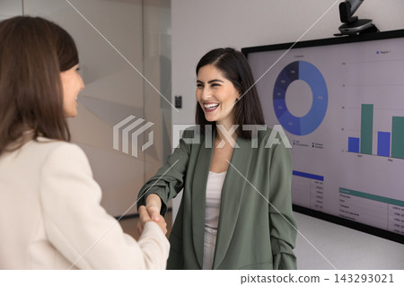 Women handshaking standing next to screen displaying charts and graphs Women handshaking standing next to screen displaying charts and graphs 143293021