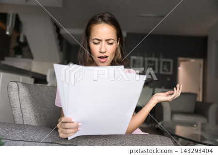 Young woman sitting on couch overwhelmed by reading paper documents Young woman sitting on couch overwhelmed by reading paper documents 143293404