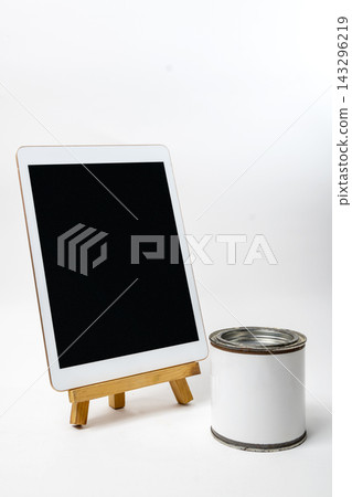 A blank tablet on a wooden easel next to a blank white paint can on a white surface, ideal for design mockups. A blank tablet on a wooden easel next to a blank white paint can on a white surface, ideal for design mockups. 143296219