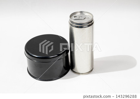 Studio shot of a round black metal container next to a tall silver beverage can on a white surface. Studio shot of a round black metal container next to a tall silver beverage can on a white surface. 143296288
