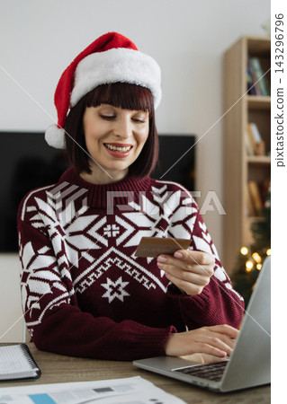 A smiling Caucasian woman wearing a Santa hat and a festive sweater is using a laptop and credit card for online shopping during the holiday season A smiling Caucasian woman wearing a Santa hat and a festive sweater is using a laptop and credit card for online shopping during the holiday season 143296796