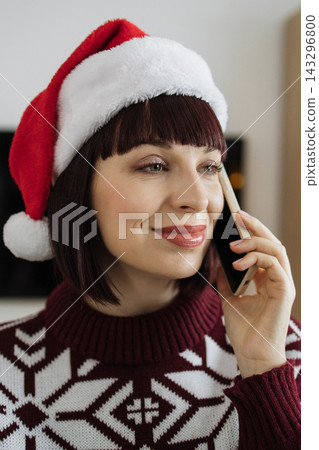 A smiling woman wearing a Santa hat and a festive sweater talks on her mobile phone during the holiday season A smiling woman wearing a Santa hat and a festive sweater talks on her mobile phone during the holiday season 143296800