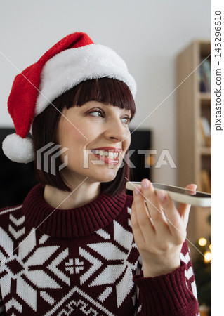 A smiling Caucasian woman in a festive sweater and Santa hat holds a smartphone to her ear, possibly recording a voice message A smiling Caucasian woman in a festive sweater and Santa hat holds a smartphone to her ear, possibly recording a voice message 143296810