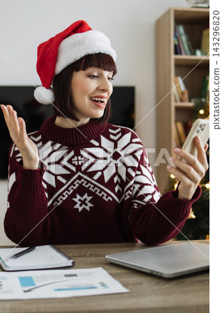 A smiling woman wearing a Santa hat and festive sweater is on a video call on her phone during the holiday season A smiling woman wearing a Santa hat and festive sweater is on a video call on her phone during the holiday season 143296820