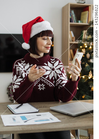 A Caucasian woman wearing a Santa hat and a festive sweater is video calling on her phone while sitting at a desk with a laptop and papers A Caucasian woman wearing a Santa hat and a festive sweater is video calling on her phone while sitting at a desk with a laptop and papers 143296822