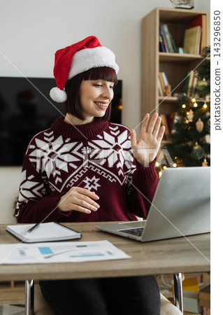 A smiling woman wearing a Santa hat and a festive sweater waves hello during a video call, with a Christmas tree in the background A smiling woman wearing a Santa hat and a festive sweater waves hello during a video call, with a Christmas tree in the background 143296850