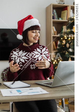 A smiling Caucasian woman wearing a Santa hat and festive sweater is engaged in a video call on her laptop with a Christmas tree in the background A smiling Caucasian woman wearing a Santa hat and festive sweater is engaged in a video call on her laptop with a Christmas tree in the background 143296851