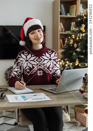 A smiling Caucasian woman wearing a Santa hat and a festive sweater works on her laptop and notebook at a desk decorated for the holidays A smiling Caucasian woman wearing a Santa hat and a festive sweater works on her laptop and notebook at a desk decorated for the holidays 143296852