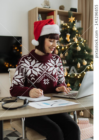 A Caucasian woman wearing a Santa hat and festive sweater works on her laptop and takes notes at a desk with a Christmas tree in the background A Caucasian woman wearing a Santa hat and festive sweater works on her laptop and takes notes at a desk with a Christmas tree in the background 143296855