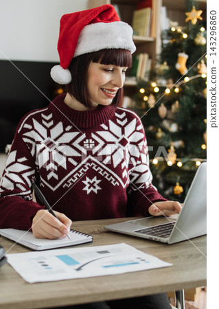 A smiling Caucasian woman wearing a Santa hat and festive sweater works on her laptop and takes notes by a decorated Christmas tree A smiling Caucasian woman wearing a Santa hat and festive sweater works on her laptop and takes notes by a decorated Christmas tree 143296860