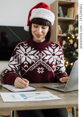 A Caucasian woman wearing a Santa hat and festive sweater works on her laptop and takes notes at a desk with Christmas decorations in the background A Caucasian woman wearing a Santa hat and festive sweater works on her laptop and takes notes at a desk with Christmas decorations in the background 143296861