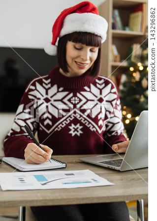 A Caucasian woman wearing a festive sweater and Santa hat works on her laptop and takes notes at a desk with holiday decorations in the background 143296862