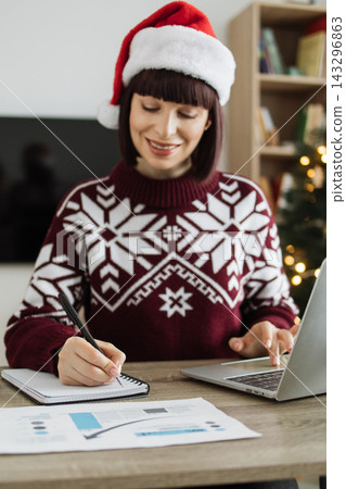 A smiling Caucasian woman wearing a Santa hat and festive sweater works on her laptop and takes notes at a desk with holiday decorations in the background 143296863