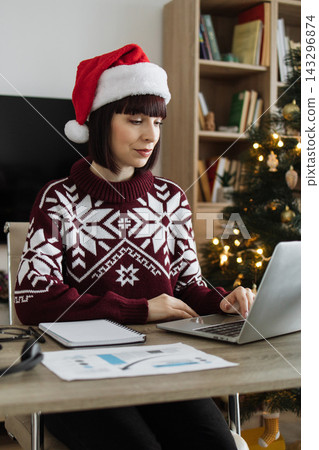 A Caucasian woman wearing a Santa hat and festive sweater works on her laptop at a desk with a Christmas tree in the background 143296874