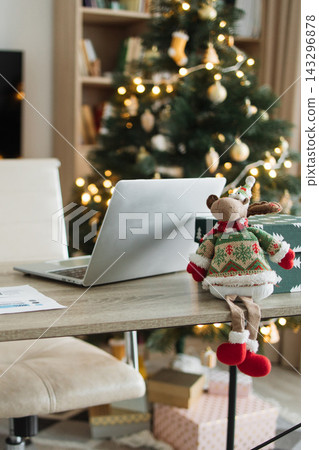 A festive office desk with a laptop, gifts, and a reindeer toy in front of a decorated Christmas tree A festive office desk with a laptop, gifts, and a reindeer toy in front of a decorated Christmas tree 143296878