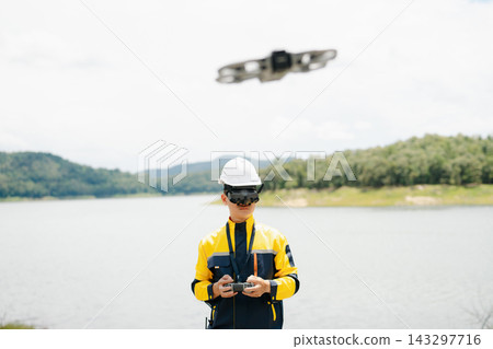 Male engineer in safety gear uses FPV goggles and a remote to pilot a drone. Conducting a high-tech aerial survey near a beautiful natural lake Male engineer in safety gear uses FPV goggles and a remote to pilot a drone. Conducting a high-tech aerial survey near a beautiful natural lake 143297716