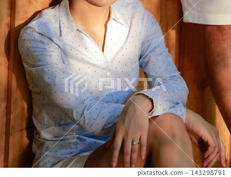 Woman sits and leans against a wooden wall wearing a light blue dotted long sleeve shirt with Sunlight soft pattern across her clothes and lap. Woman sits and leans against a wooden wall wearing a light blue dotted long sleeve shirt with Sunlight soft pattern across her clothes and lap. 143298791