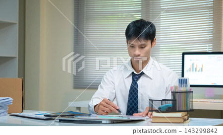 Businessman Working at Office Desk Reviewing Financial Charts and Documents with Pen Businessman Working at Office Desk Reviewing Financial Charts and Documents with Pen 143299842