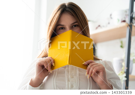 Young woman holding a yellow notebook over her face in a bright home interior. Personal diary privacy creative learning and stationery Young woman holding a yellow notebook over her face in a bright home interior. Personal diary privacy creative learning and stationery 143301339