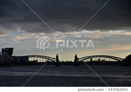 Silhouetted steel arch bridge spanning wide river under dramatic cloudy sky at dusk with city skyline in background urban infrastructure Silhouetted steel arch bridge spanning wide river under dramatic cloudy sky at dusk with city skyline in background urban infrastructure 143302042