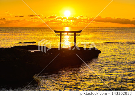 A winter scene on Oarai Beach: the rising sun over the silhouette of the Kamiiso Torii gate, the golden-hued sea, and the sunlit road. A winter scene on Oarai Beach: the rising sun over the silhouette of the Kamiiso Torii gate, the golden-hued sea, and the sunlit road. 143302933