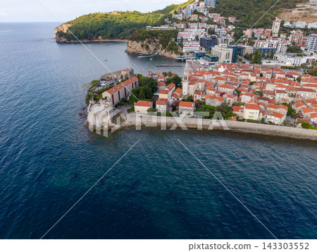 Budva Montenegro coastline frames historic walled town and calm bay. Aerial view includes terracotta rooftops, stone fortifications, church spire and modern buildings on green slopes. Budva Montenegro coastline frames historic walled town and calm bay. Aerial view includes terracotta rooftops, stone fortifications, church spire and modern buildings on green slopes. 143303552