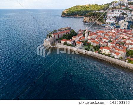 Budva Montenegro historic center occupies walled peninsula along Adriatic coast. Stone buildings with terracotta roofs cluster around church tower, with modern resorts along nearby shore. Budva Montenegro historic center occupies walled peninsula along Adriatic coast. Stone buildings with terracotta roofs cluster around church tower, with modern resorts along nearby shore. 143303553