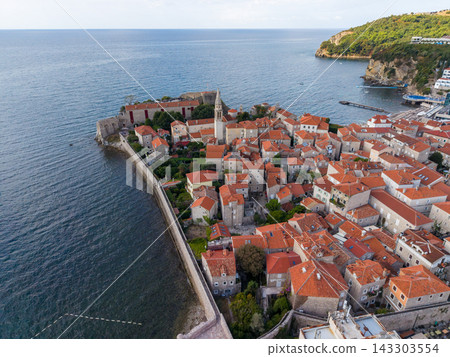 Budva Montenegro old town sits within stone coastal walls beside blue Adriatic water. Dense historic buildings with red tile roofs cluster around slender church bell tower on peninsula. Budva Montenegro old town sits within stone coastal walls beside blue Adriatic water. Dense historic buildings with red tile roofs cluster around slender church bell tower on peninsula. 143303554