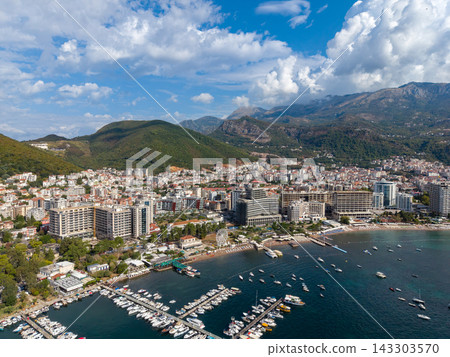 Budva Montenegro appears in sweeping coastal view, with harbor boats, beachside piers and buildings along shore. Adriatic resort town sits beneath green mountain slopes and cloud filled sky. 143303570