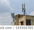 Weathered building rooftop hosts two distinct telecommunications antenna arrays against bright blue sky with scattered clouds 143303765