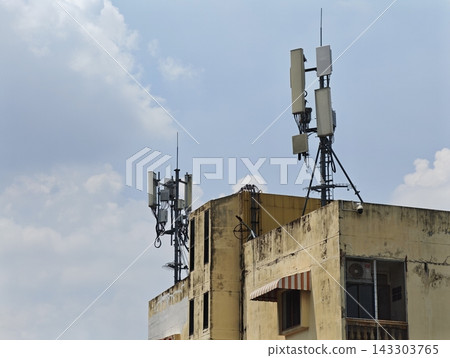 Weathered building rooftop hosts two distinct telecommunications antenna arrays against bright blue sky with scattered clouds Weathered building rooftop hosts two distinct telecommunications antenna arrays against bright blue sky with scattered clouds 143303765