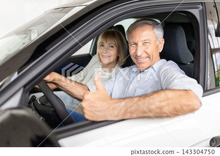 Smiling senior couple sitting inside car at dealership showroom, showing thumb up Smiling senior couple sitting inside car at dealership showroom, showing thumb up 143304750