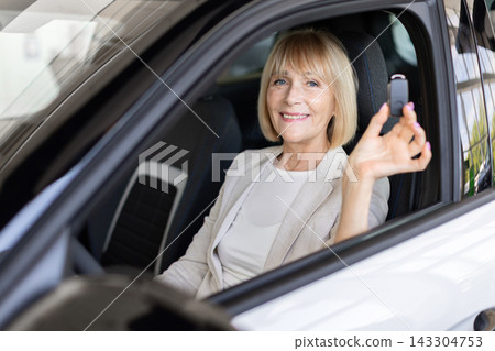 Smiling senior woman sitting inside vehicle at dealership showroom, displays car key Smiling senior woman sitting inside vehicle at dealership showroom, displays car key 143304753