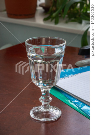 Clear water in a stemmed glass on a desk beside a notebook and pen. Drinking water and hydration. Everyday refreshment and healthy routine. Desk-side refreshment for work and study. Clear water in a stemmed glass on a desk beside a notebook and pen. Drinking water and hydration. Everyday refreshment and healthy routine. Desk-side refreshment for work and study. 143305530