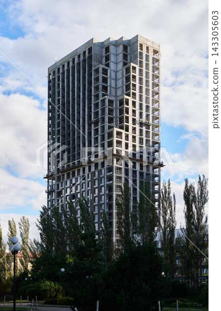 Under Construction Highrise Tower with exposed concrete frame and grid of windows rising above trees and street lamps against bright blue sky with scattered clouds and city backdrop Under Construction Highrise Tower with exposed concrete frame and grid of windows rising above trees and street lamps against bright blue sky with scattered clouds and city backdrop 143305603