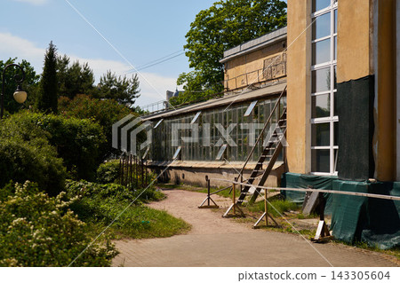 Yellow building facade with glass greenhouse extension, wooden staircase, lush garden and greenery under bright blue sky in urban setting, historic rustic exterior bathed in sunlight Yellow building facade with glass greenhouse extension, wooden staircase, lush garden and greenery under bright blue sky in urban setting, historic rustic exterior bathed in sunlight 143305604