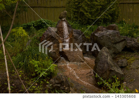 Rock garden fountain with flowing water cascading over stacked stones, surrounded by vibrant greenery and bamboo fence within tranquil outdoor garden with sunlit stone pathway Rock garden fountain with flowing water cascading over stacked stones, surrounded by vibrant greenery and bamboo fence within tranquil outdoor garden with sunlit stone pathway 143305608
