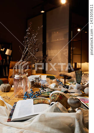 Cozy indoor still life with open magazine, pink glass vase holding dried branches, blue berries, ceramics, and textiles on wooden table under warm ambient lighting 143305611