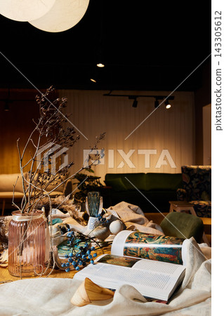 Dining table centerpiece with pink ribbed glass vase, dried branches, and opened magazine under warm indoor lighting with soft shadows for home decor Dining table centerpiece with pink ribbed glass vase, dried branches, and opened magazine under warm indoor lighting with soft shadows for home decor 143305612